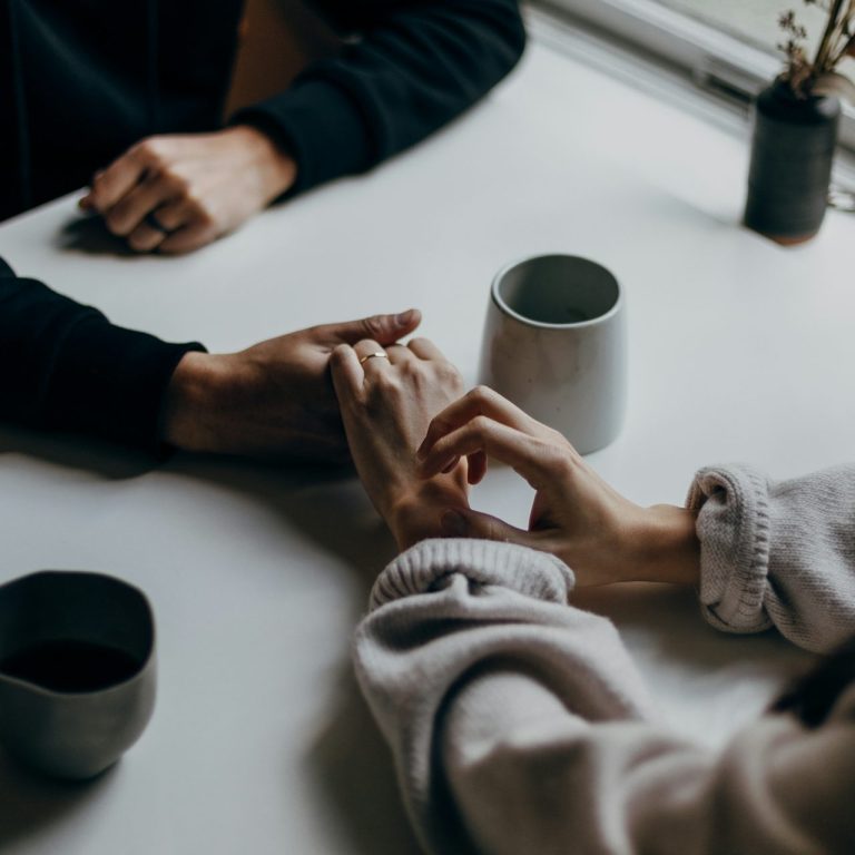 Zwei Hände halten sich über einem Tisch mit Tassen und einer kleinen Pflanze.
