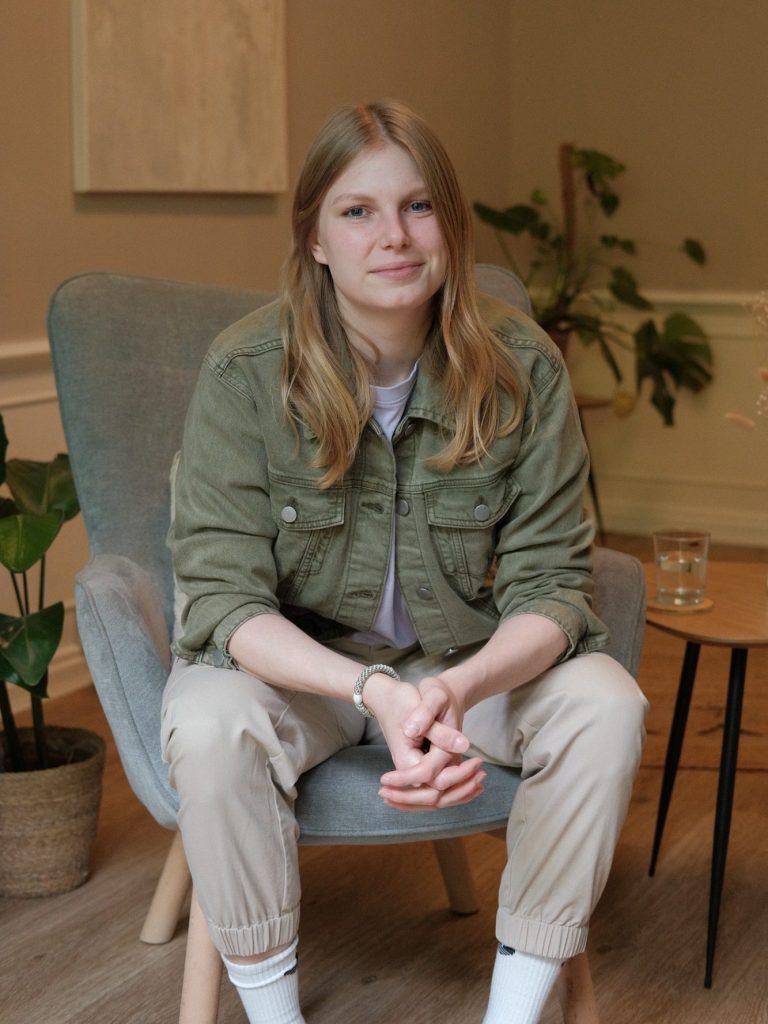 Beraterin im Raum Das Bild zeigt die Beraterin Janine Kröger im Praxisraum. Eine junge Frau mit langen Haaren sitzt in einem Sessel, umgeben von Pflanzen.