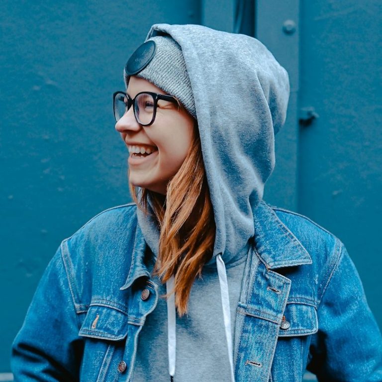 Junge Frau mit Brille, Kapuzenpullover und Jeansjacke lächelt vor blauer Wand.