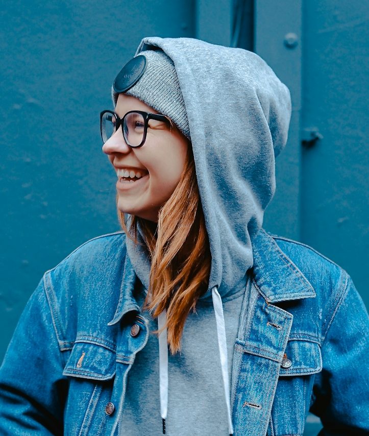 Fröhliche Frau Junge Frau mit Brille, Hoodie und Jeansjacke, lächelt vor einer blauen Wand.