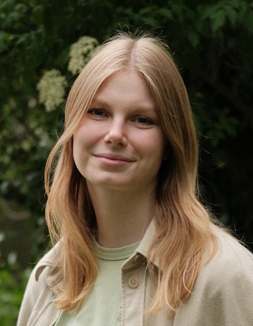Portrait Das Bild zeigt ein Portrait der Beraterin Janine Kröger, eine junge Frau mit langen, blonden Haaren, lächelnd vor grünen Pflanzen.