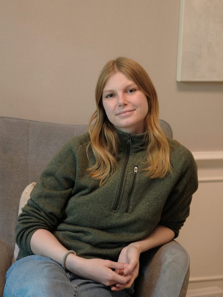 Die Beraterin Janine Kröger, eine junge Frau mit langen, blonden Haaren sitzt entspannt auf einem Sessel und lächelt in die Kamera.